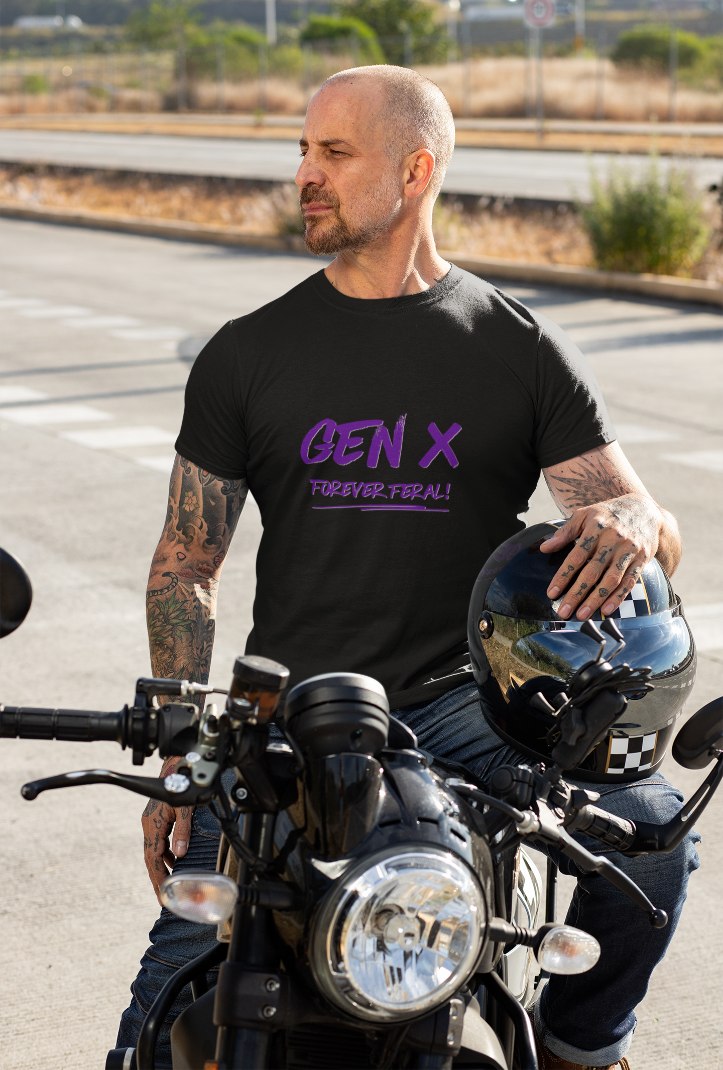 An attractive Gen X male sits on his motorcycle wearing a black organic cotton unisex t-shirt with the words Gen X Forever Feral across the chest in purple.