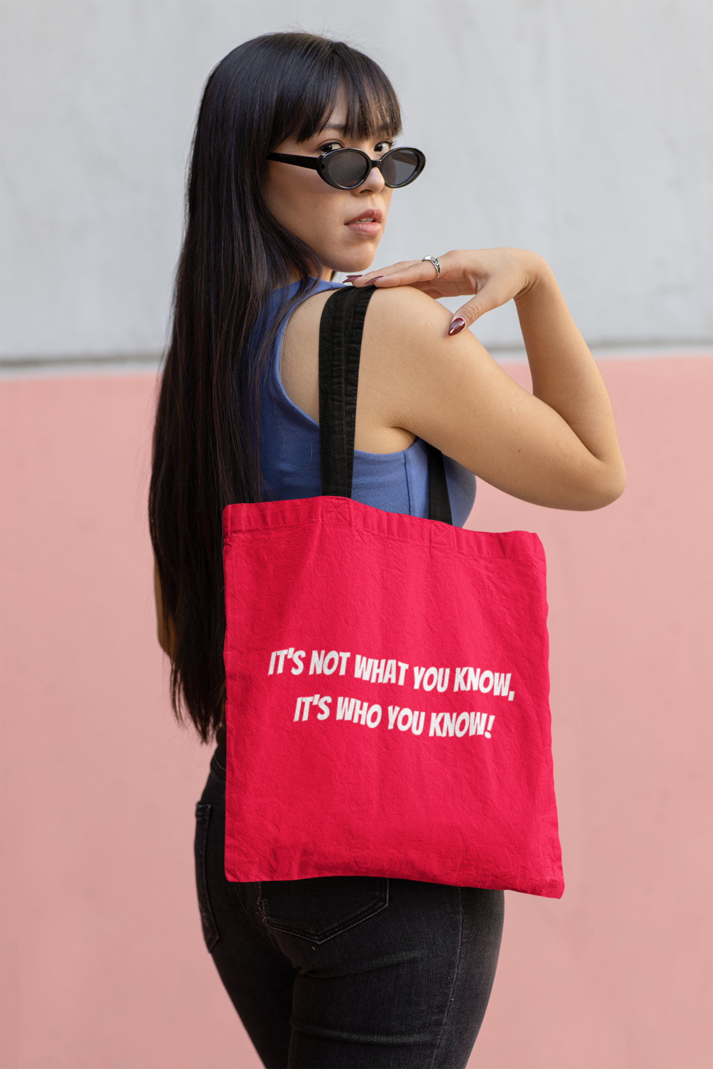 A beautiful Asian woman wearing a sleeveless top is looking over her shoulder toward the viewer. She is carrying a torch red canvas shopping tote with black handles. On the side of the tote reads It's not what you know, It's who you know! in white letters.