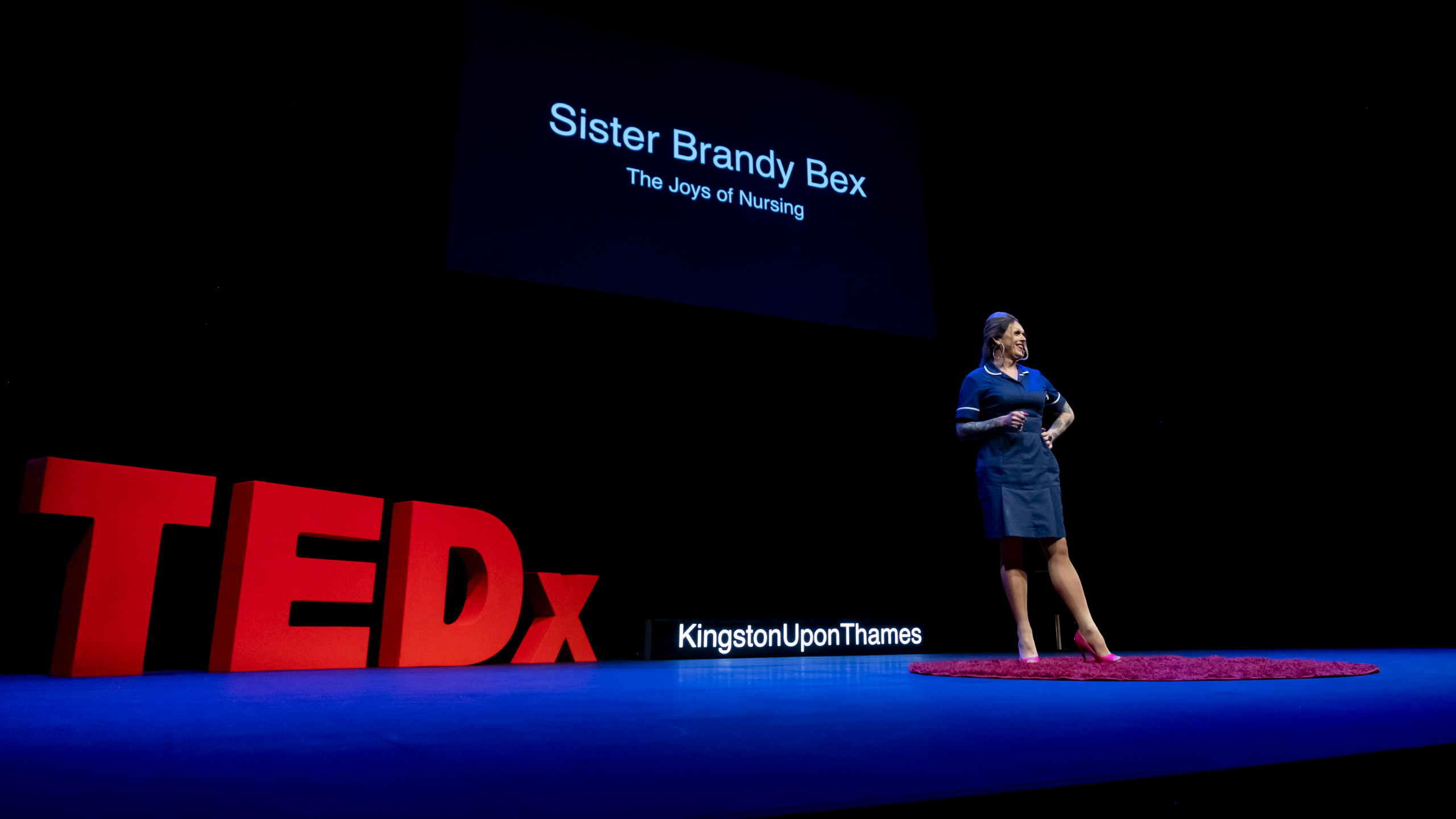 Load video: Sister Brandy Bex on stage at TEDx Kingston Upon Thames. Brandy is wearing her Band 6 navy nursing uniform and is standing on the iconic red circular rug. The large TEDx letter are visible in bright red at the rear of the stage.